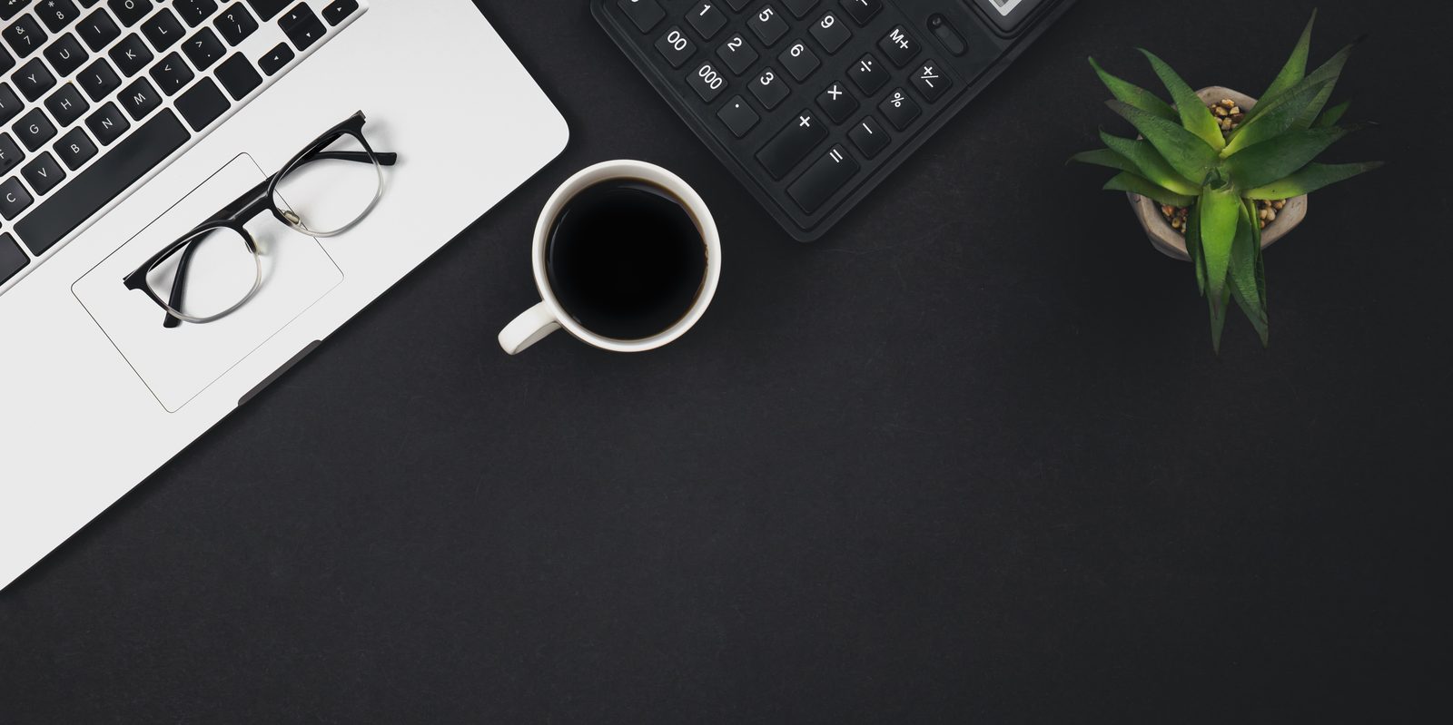 Escritorio de asesoría sobre fondo negro con portátil, calculadora, café y gafas, vista cenital